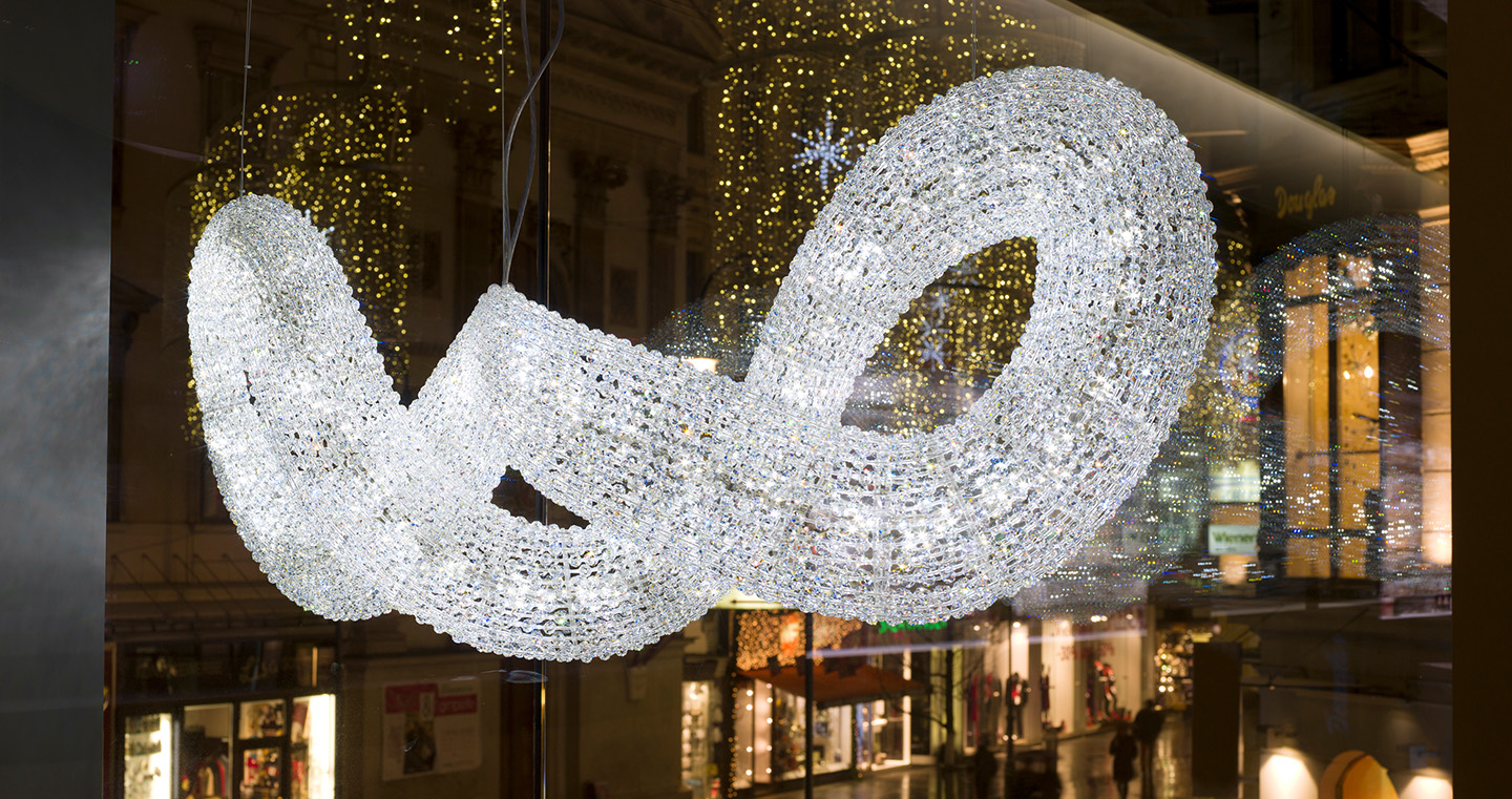 Schweifende Lichtskulptur von Yves Béhar: Eine geschwungene, funkelnde Installation aus tausenden Kristallen, die wie eine schwebende Schlaufe wirkt. Im Hintergrund festliche Lichter der Stadt.