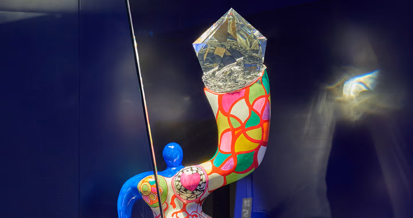 Farbintensive Skulptur von Niki de Saint Phalle: Eine bunte, organisch geformte Figur mit kräftigen Mustern in Blau, Rot, Grün und Gelb, die einen übergroßen Kristall auf einer ausgestreckten Armform trägt.