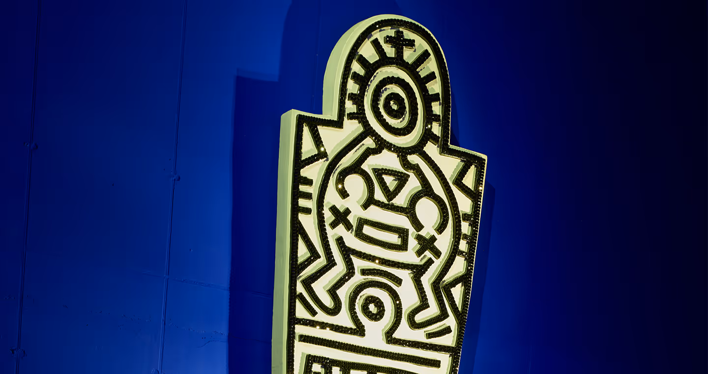 Wandinstallation in mit einem typischen Keith-Haring-Motiv: eine stilisierte, grafische Figur mit schwarzen Kristallen auf weißem Grund mit Symbolen und Mustern. Das Werk leuchtet auffällig vor der blauen Wand.