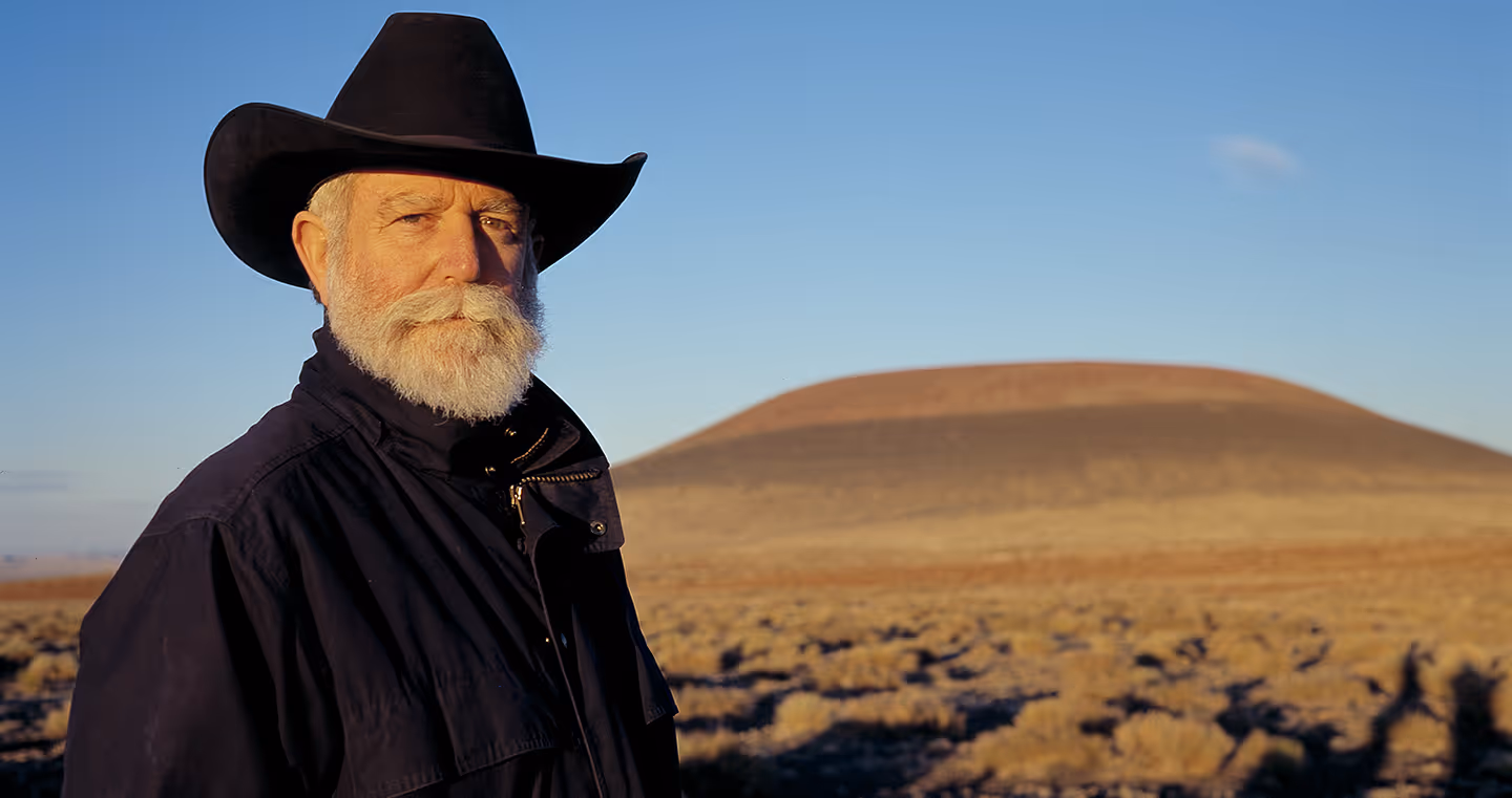 James Turrell, US-amerikanischer Künstler, im Porträt mit Hut, Brille und weißem Bart, vor dunklem Dünenlandschaft.