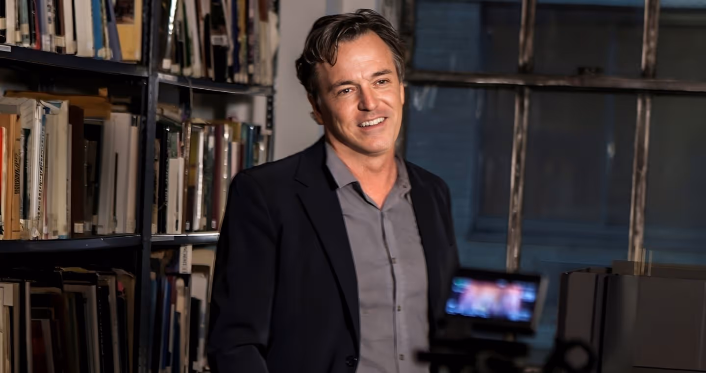 Derek McLane, US-amerikanischer Bühnenbildner, im Porträt mit schwarzer Jacke, stehend vor Bücherregalen.