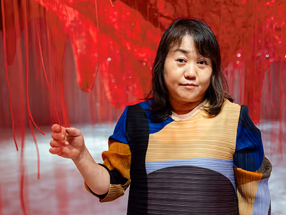 Chiharu Shiota, japanische Künstlerin, 2025 in farbenfrohem Kleid, stehend inmitten ihrer roten Fadeninstallation.