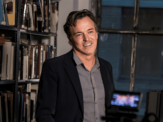 Derek McLane, US-amerikanischer Bühnenbildner, im Porträt mit schwarzer Jacke, stehend vor Bücherregalen.