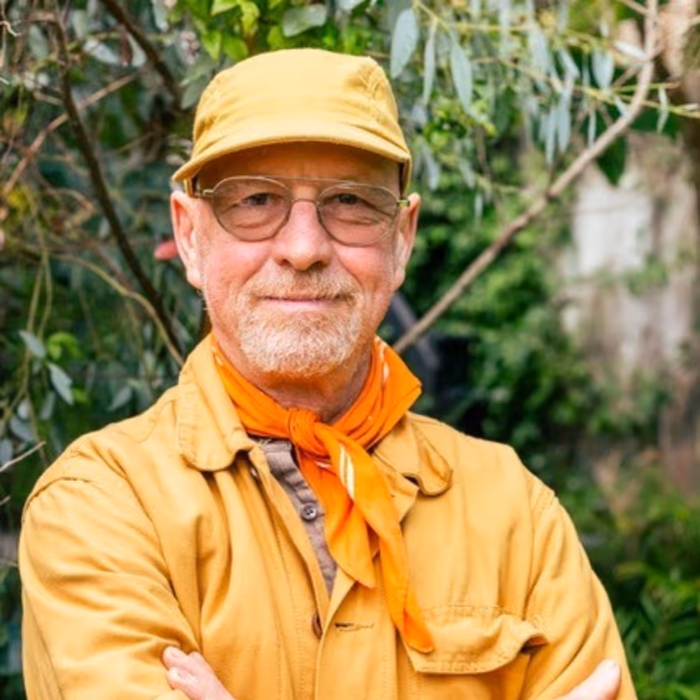 Portrait of a person wearing glasses, a light-colored cap, and a yellow-ochre jacket, standing in front of a green, overgrown background with their arms crossed in front of their upper body; an orange scarf is tied around their neck.