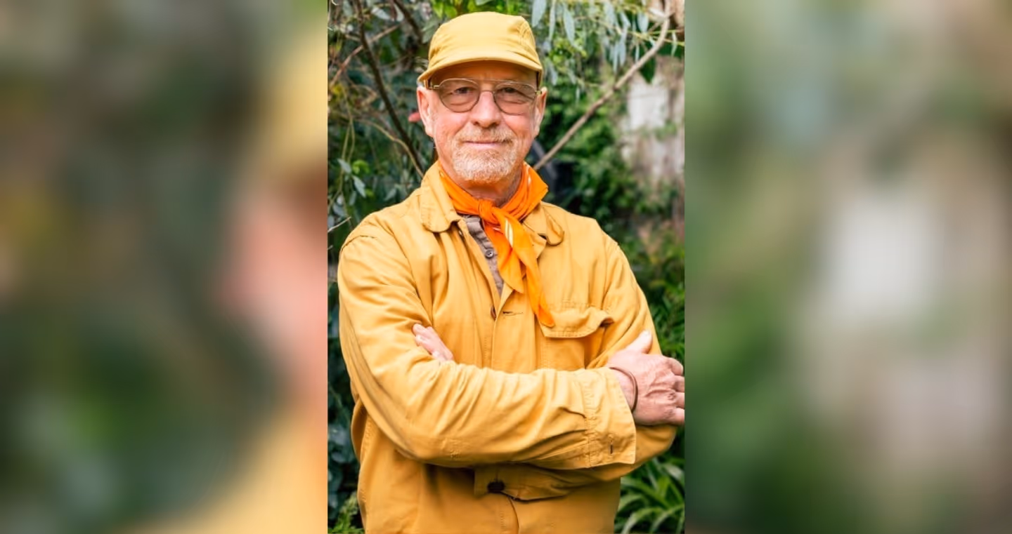 Portrait of a person wearing glasses, a light-colored cap, and a yellow-ochre jacket, standing in front of a green, overgrown background with their arms crossed in front of their upper body; an orange scarf is tied around their neck.