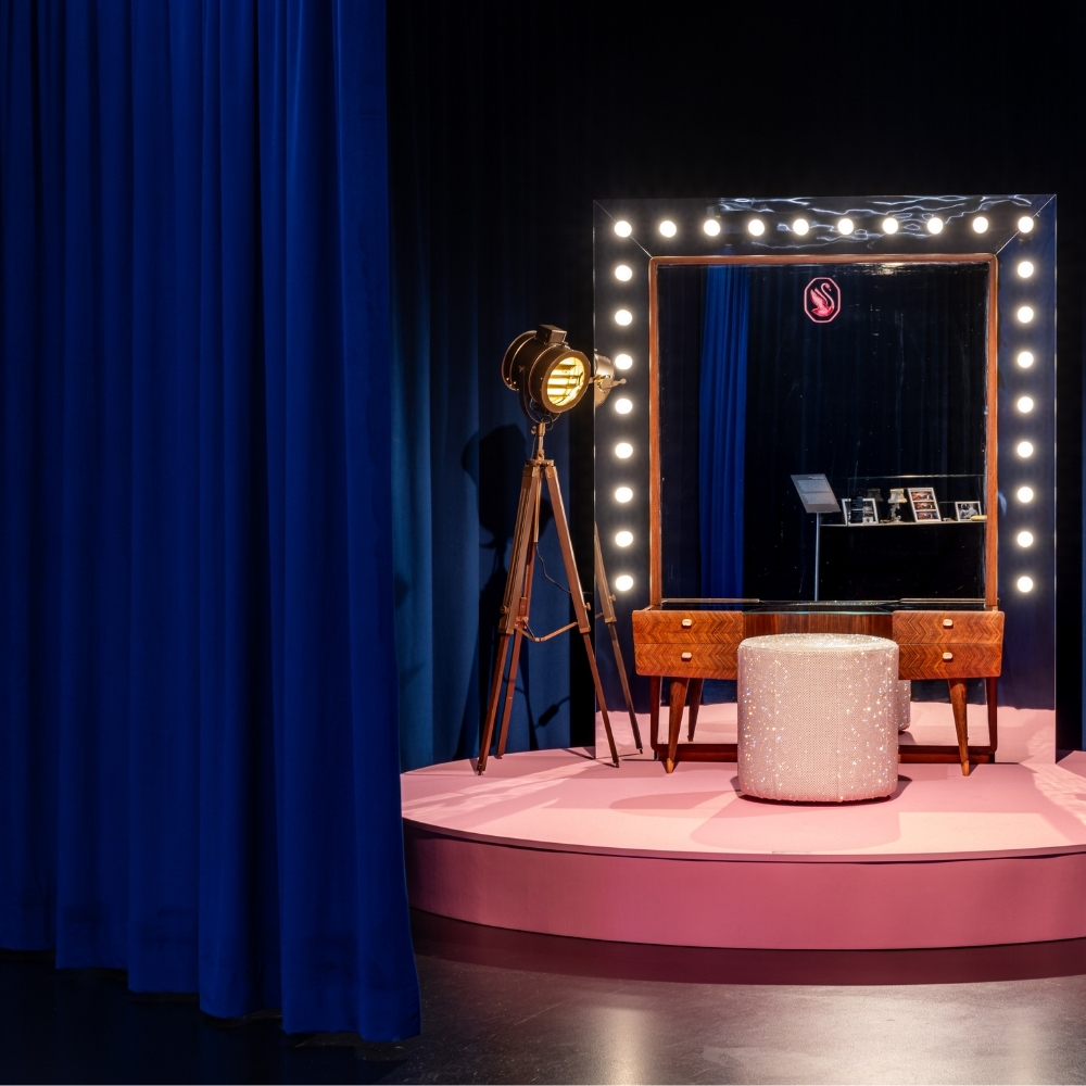 Eleganter Schminkplatz auf rosa Bühne mit beleuchtetem Spiegel, funkelndem Hocker und Studiolampe, eingerahmt von dunkelblauen Vorhängen.