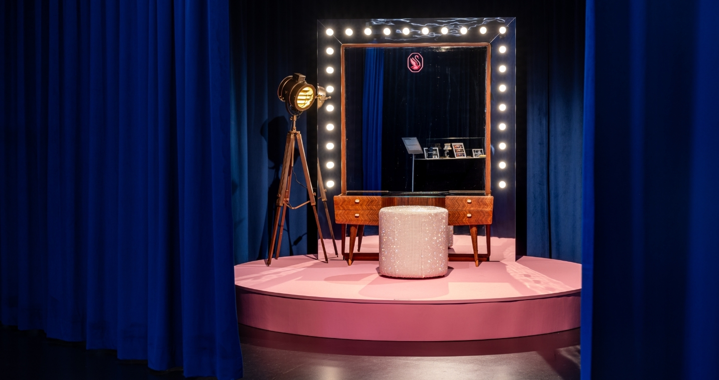Eleganter Schminkplatz auf rosa Bühne mit beleuchtetem Spiegel, funkelndem Hocker und Studiolampe, eingerahmt von dunkelblauen Vorhängen.
