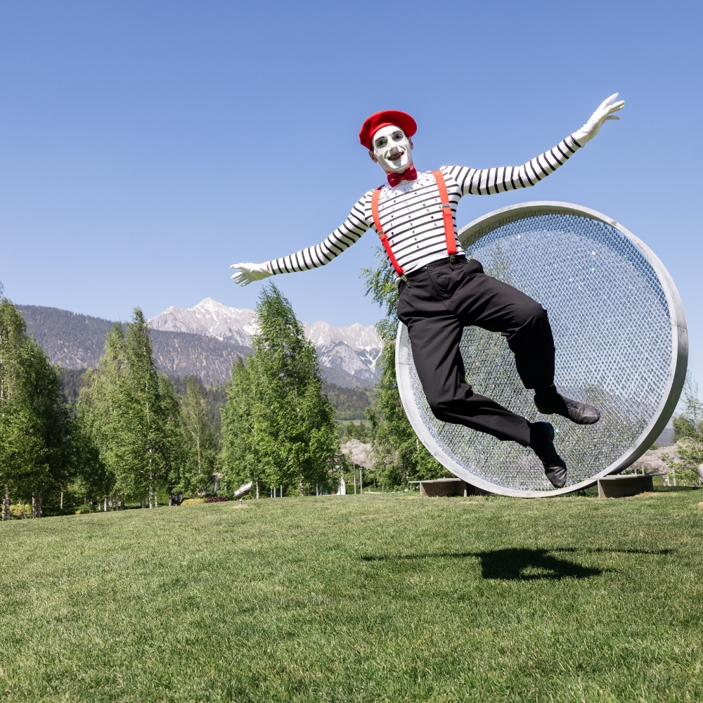 Pantomime in gestreiftem Shirt, roter Mütze und Hosenträgern springt im Park der Swarovski Kristallwelten mit Bäumen und Bergen im Hintergrund.