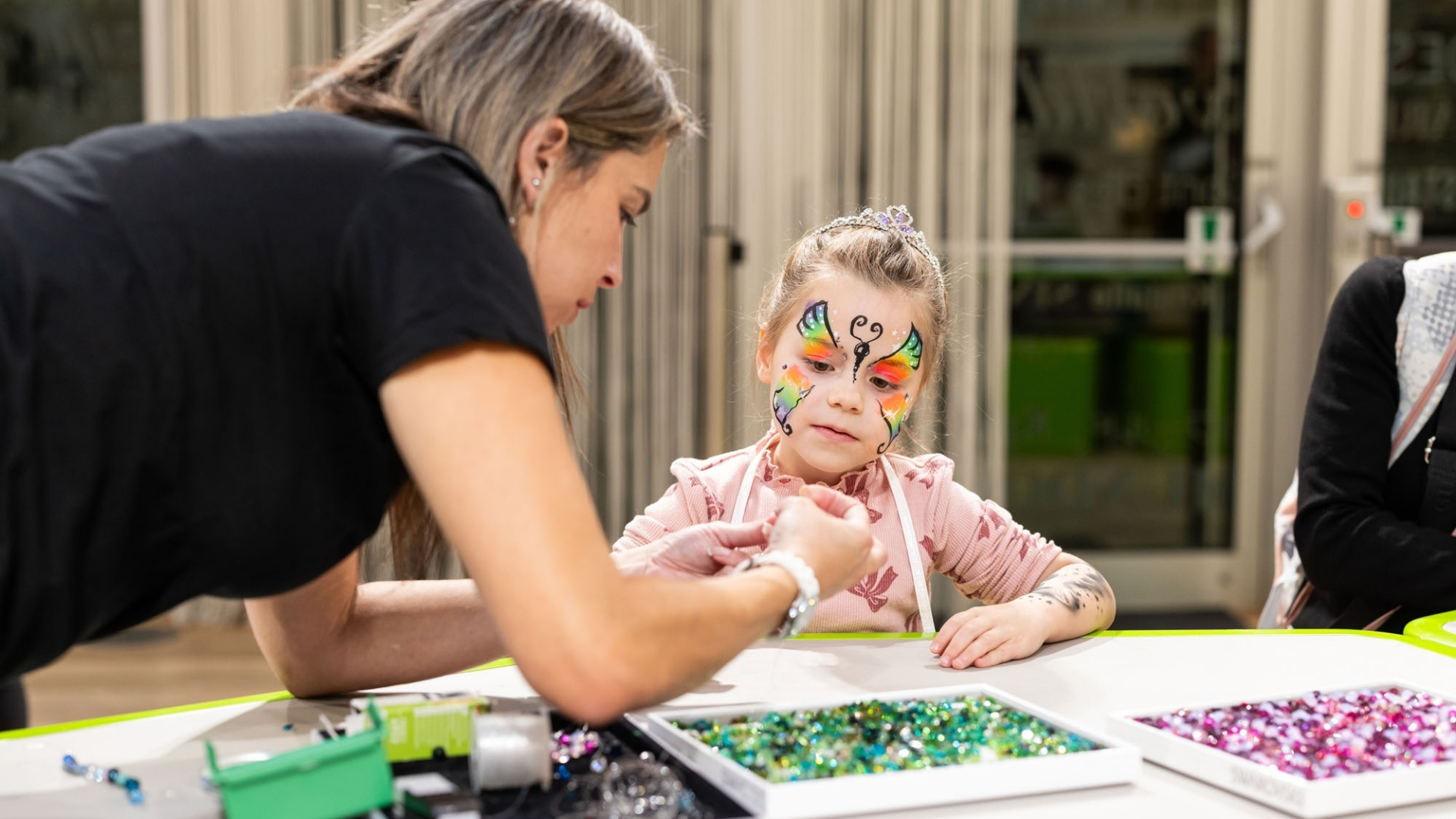 Kind mit bunter Gesichtsbemalung sitzt an einem Tisch, während eine Person Kristalle für einen kreativen Workshop vorbereitet.