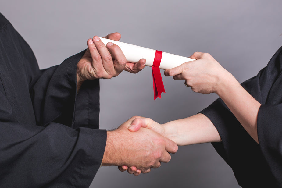 Two people exchanging a rolled diploma with a red ribbon over a handshake