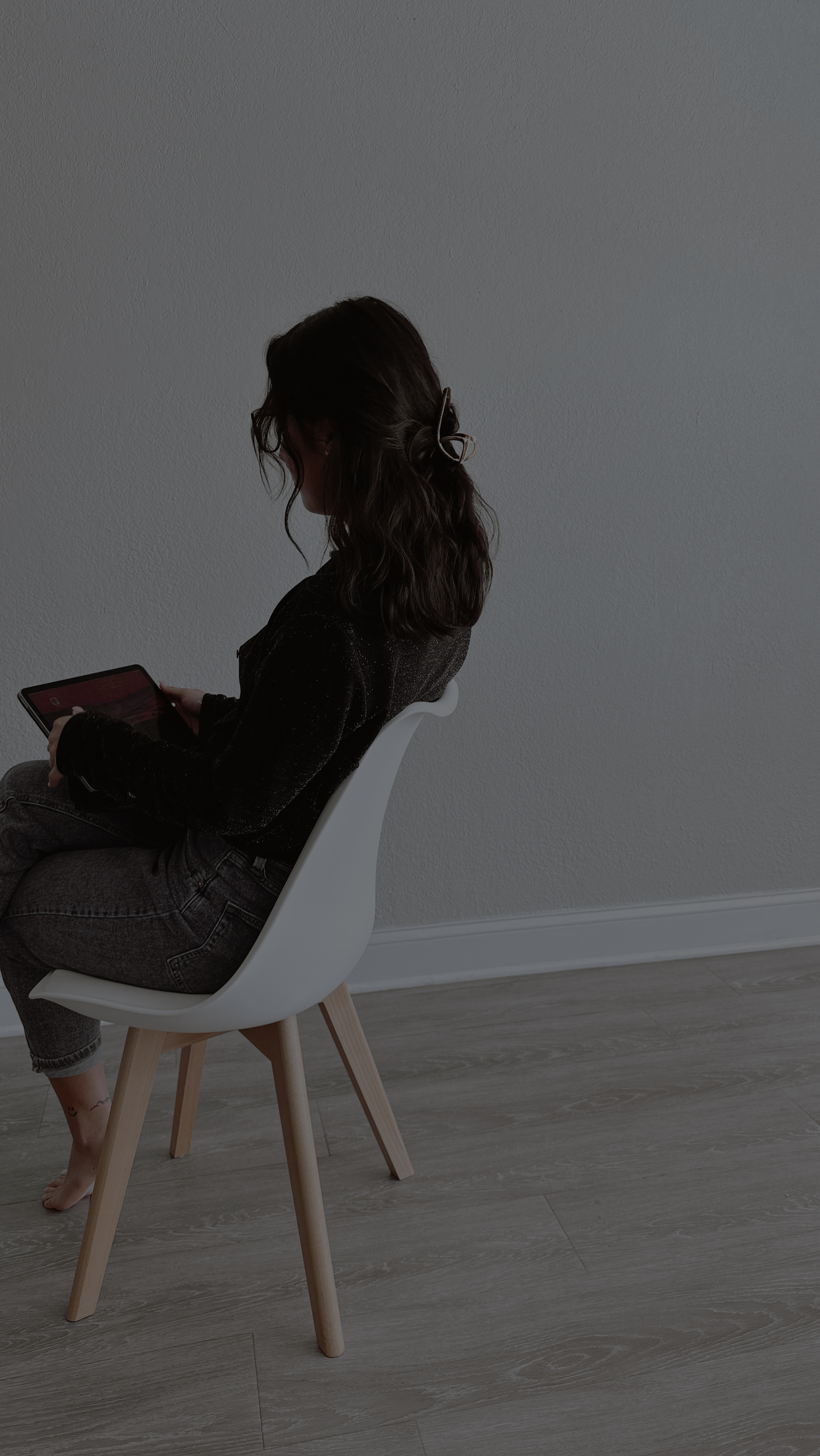 Symbolbild zu ‘Wie einsam macht das moderne Leben’: junge Frau sitzt allein auf einem Stuhl in einem leeren Raum und blickt auf ein Tablet, stellvertretend für digitale Vernetzung ohne persönliche Nähe.