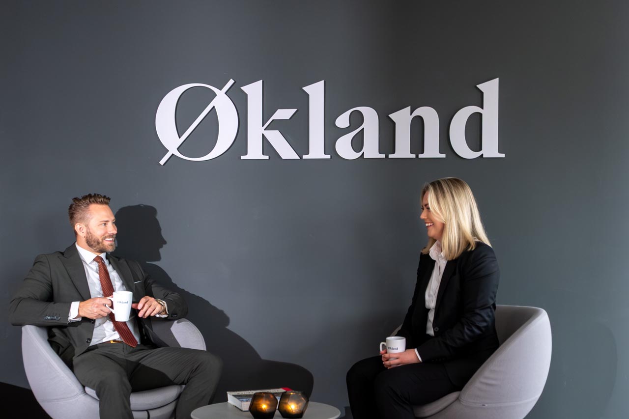 to ansatte i advokatfirmaet økland som sitter i stoler foran en vegg med et stort skilt der det står Økland