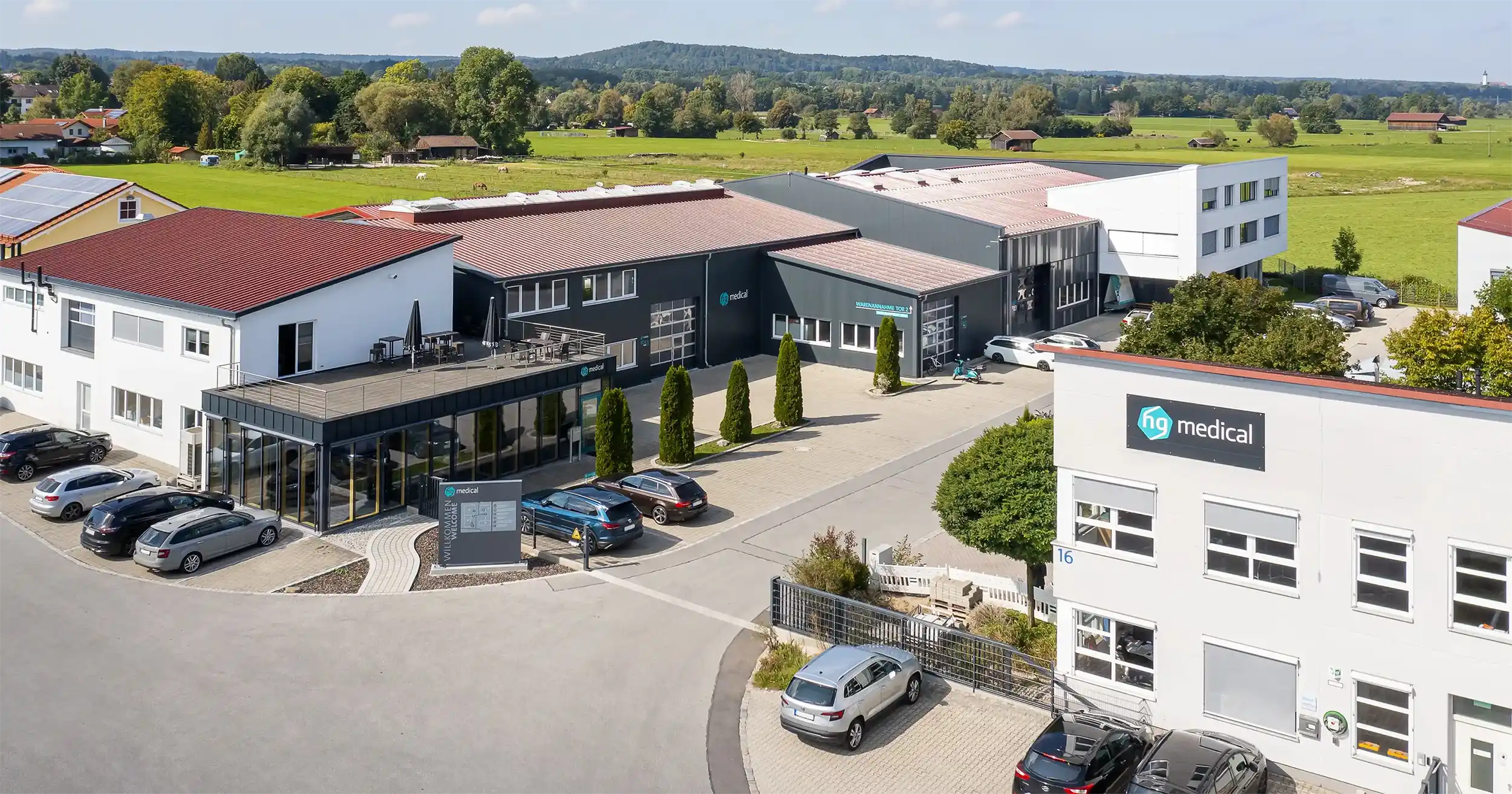 Aerial view of the hg medical company buildings and parking lot, surrounded by green fields and trees on a sunny day in a rural area.