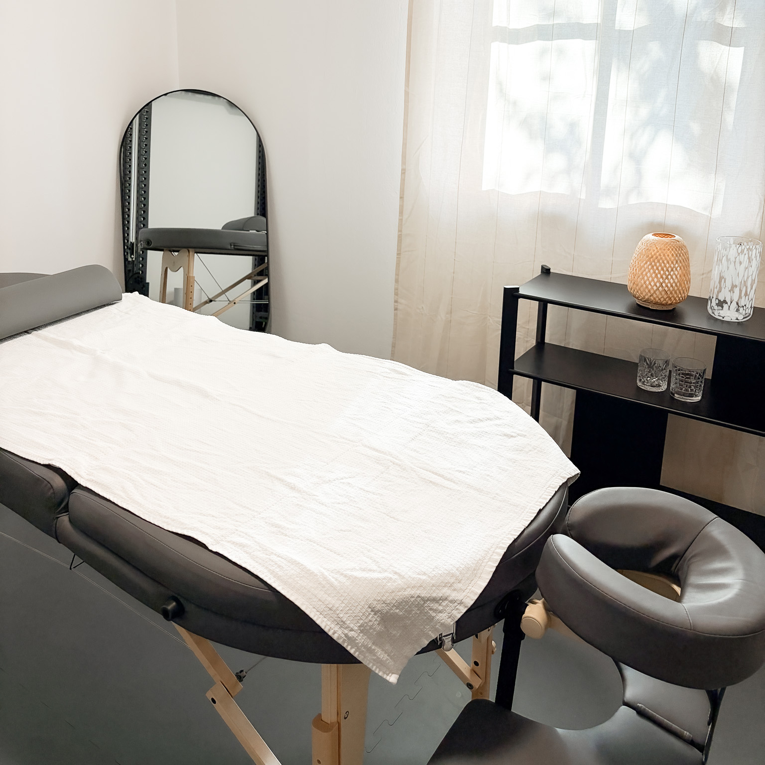 Minimalist massage room with a black massage table covered by a white towel, a face cradle, a standing mirror, and a shelf with decorative items by a window with sheer curtains.