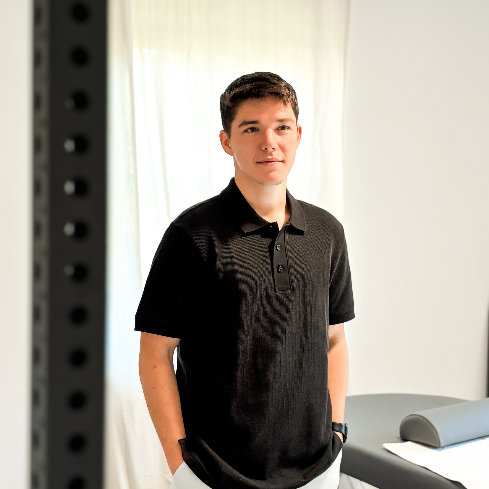 Young man with short dark hair wearing a black polo shirt and light pants, standing indoors near a therapy or massage table.