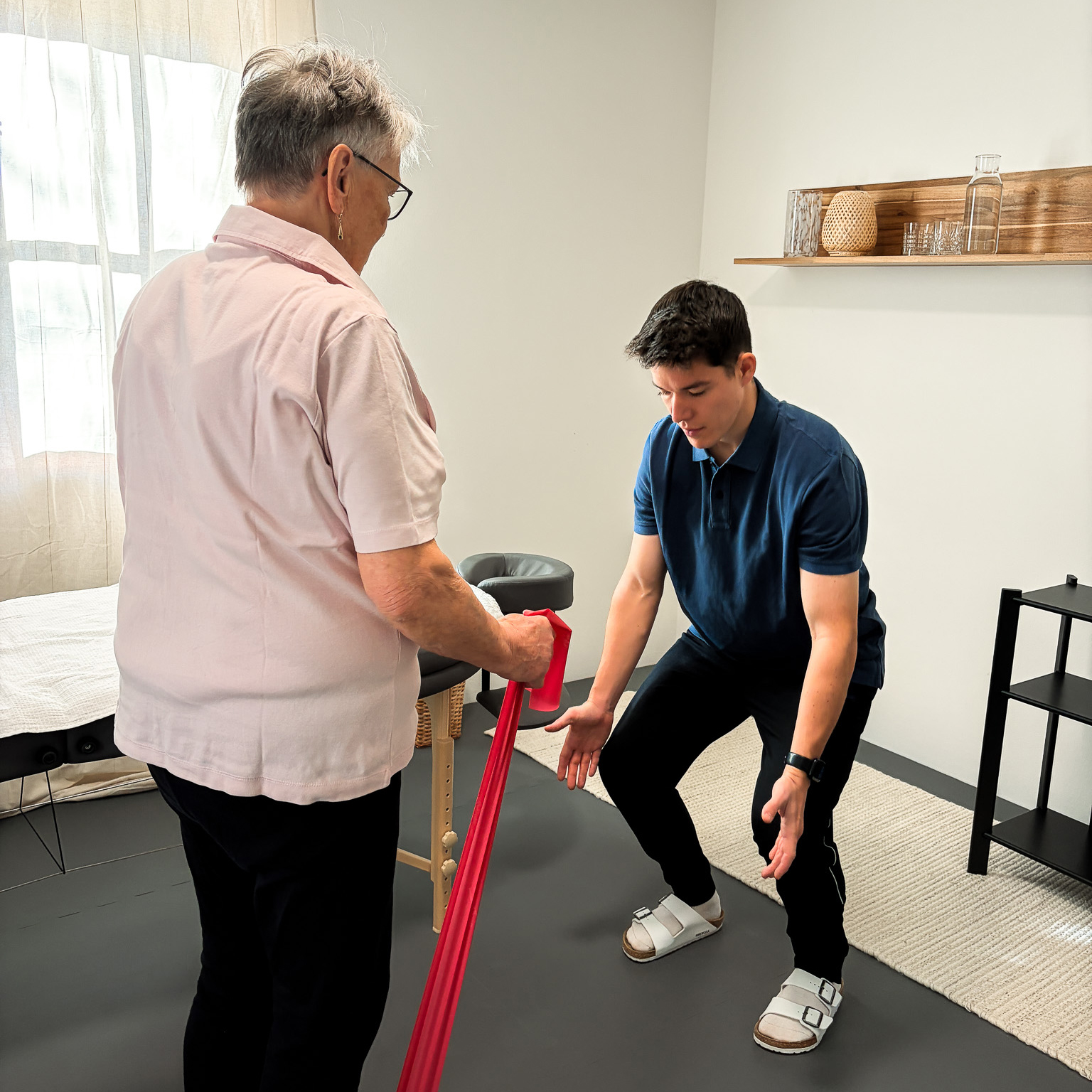 Young male physical therapist assisting an elderly woman using a red resistance band in a bright therapy room.