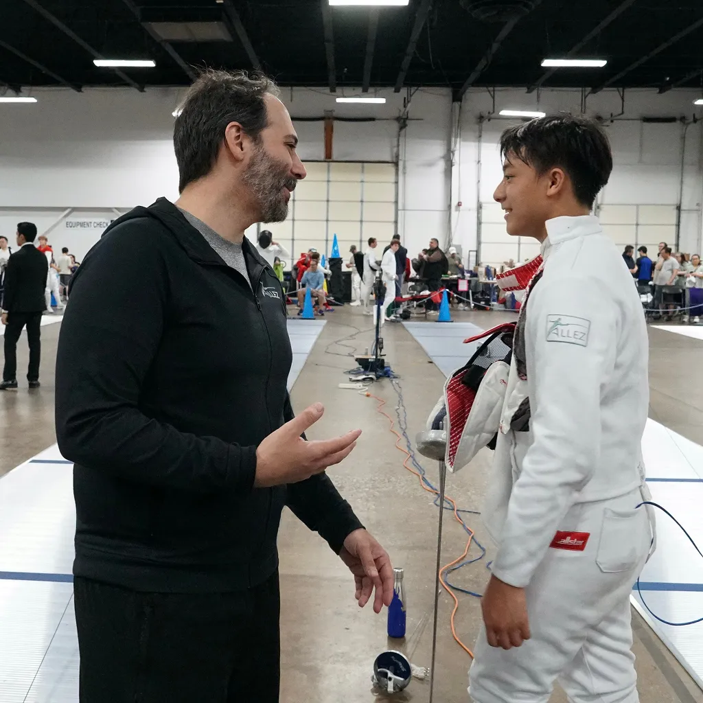 Coach from Allez Fencing Club talks with a young épée fencer during a fencing competition, highlighting mentorship and support at a tournament setting.