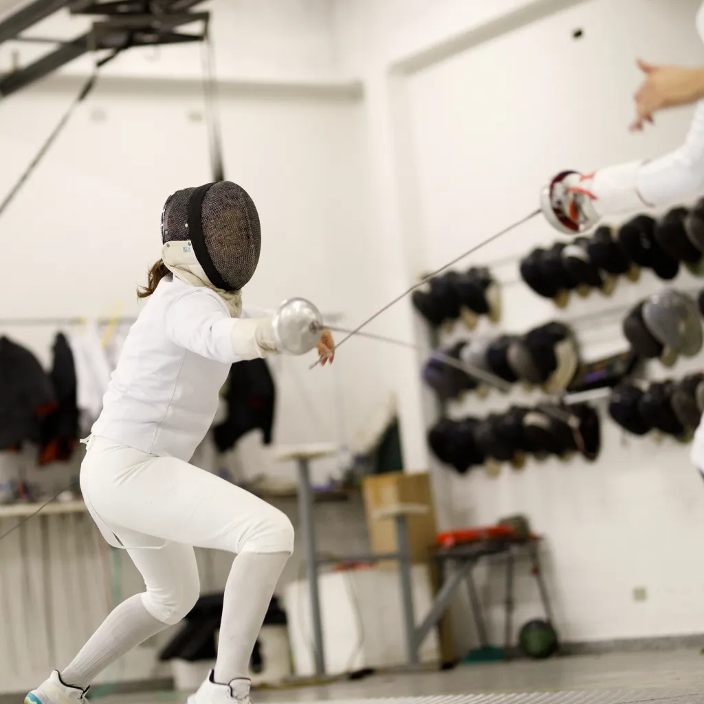 Female épée fencer performing a deep lunge during a bout at Allez Fencing Club in Watertown, MA, with fencing masks and gear visible in the background.
