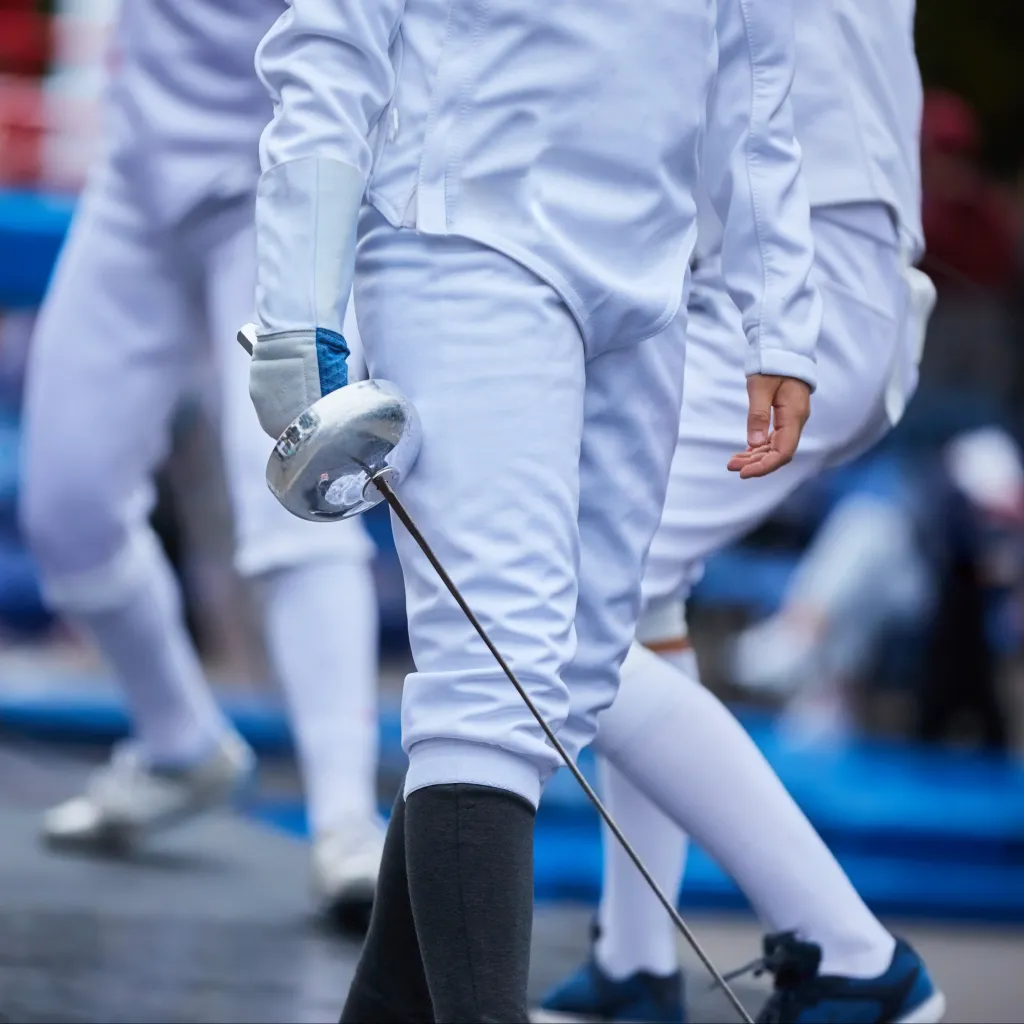 Close-up of an Allez Fencing Club épée fencer holding their weapon during a youth fencing competition, with other fencers in the background.