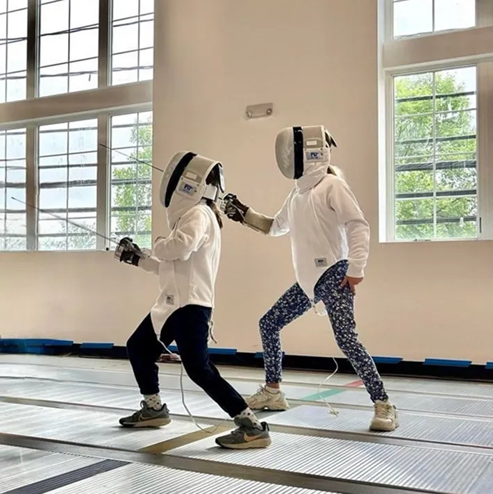 Two young fencers practicing footwork and bladework in an épée class at Allez Fencing Club in Watertown, MA, inside a bright, modern fencing facility.