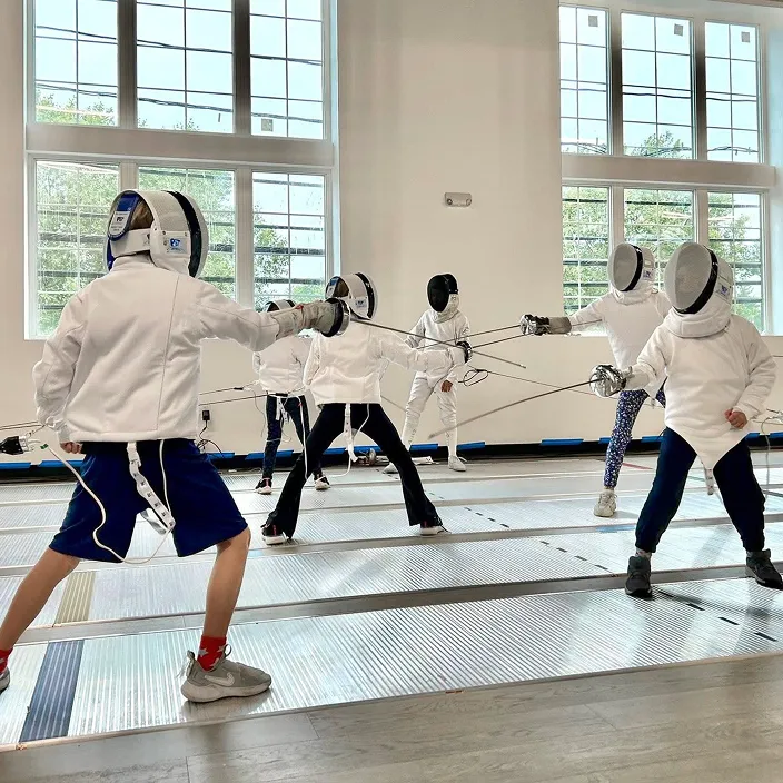 Young fencers in full fencing equipment holding épée swords and masks during a beginner fencing class at a training club.