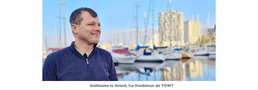Portrait de Guillaume le Grand, co-fondateur de TOWT (Transport à la voile), debout sur un quai de port de plaisance. Il regarde vers la gauche en souriant, vêtu d’un pull zippé bleu marine. En arrière-plan, des voiliers sont amarrés dans une marina avec des immeubles modernes visibles sous un ciel dégagé. Légende en dessous : « Guillaume le Grand, Co-fondateur de TOWT ».