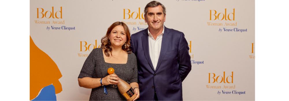 Amélie Guicheney pose avec un homme en costume sur le tapis photo du Bold Woman Award by Veuve Clicquot. Elle tient un trophée en forme de bouteille de champagne en bois, sourire aux lèvres. En arrière-plan, un mur blanc imprimé du logo « Bold Woman Award by Veuve Clicquot » en orange et bleu.