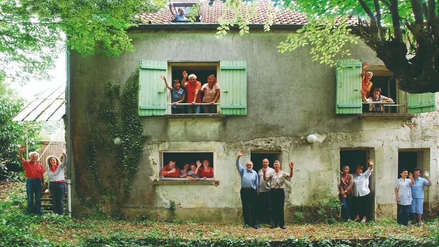 Groupe de personnes souriantes saluant depuis les fenêtres et l’entrée d’une grande maison ancienne.