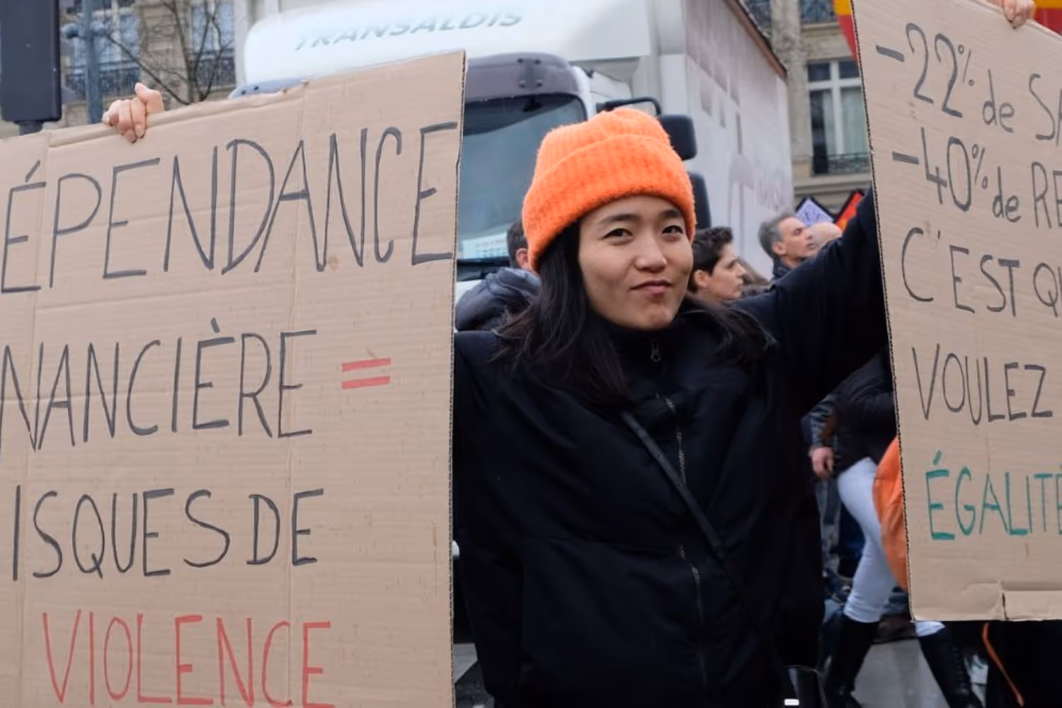 Femme manifestant avec deux pancartes en carton dénonçant les inégalités économiques et les violences liées à la dépendance financière.