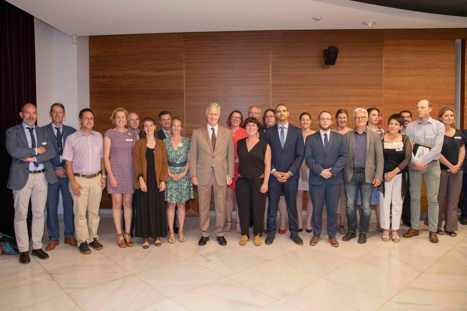 Photo de groupe lors d’un événement officiel, avec le roi Philippe de Belgique au centre, entouré d’une vingtaine de personnes souriantes