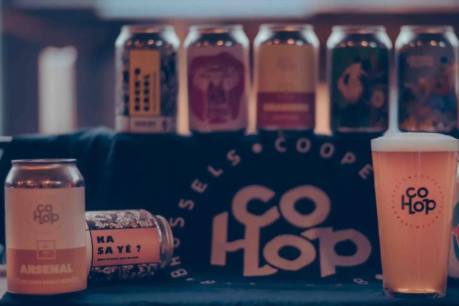 Een uitstalling van ambachtelijke bieren geproduceerd door de CoHop coöperatie in Brussel. Op de voorgrond: twee blikjes (waaronder één met de naam “Arsenal”) en een glas gevuld met pils met het CoHop-logo. Op de achtergrond: een rij van zes blikjes met verschillende kleurrijke grafische ontwerpen, geplaatst op een zwart doek met de vermelding “Brussels Coop CoHop”.