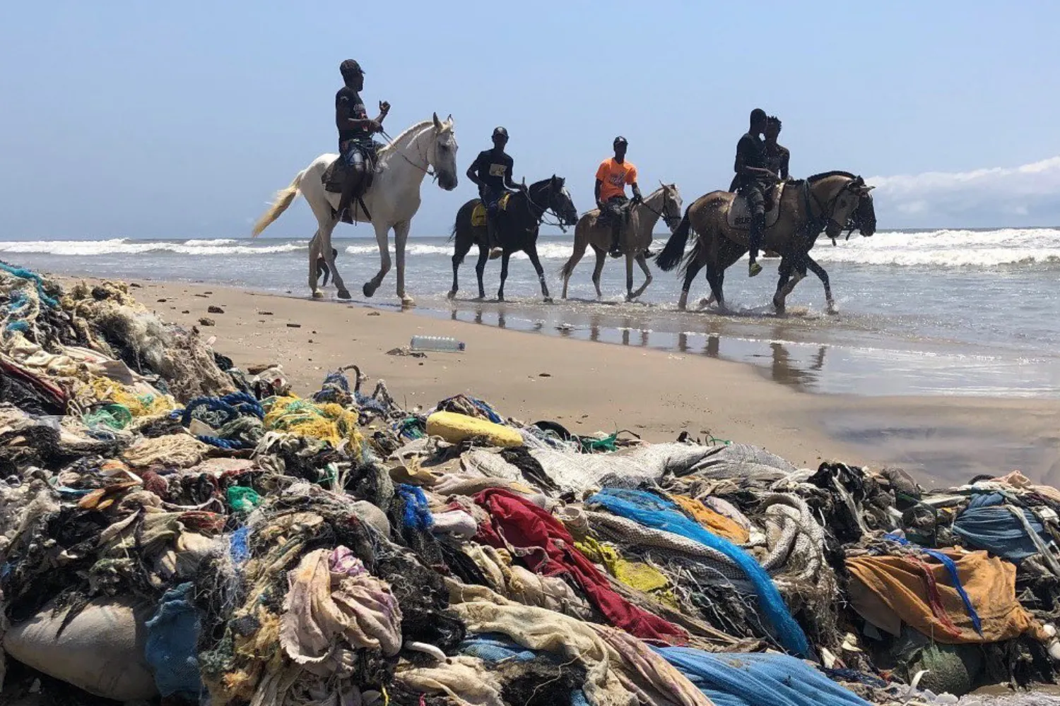Déchets textiles amoncelés sur une plage avec des cavaliers à cheval au bord de l’eau.