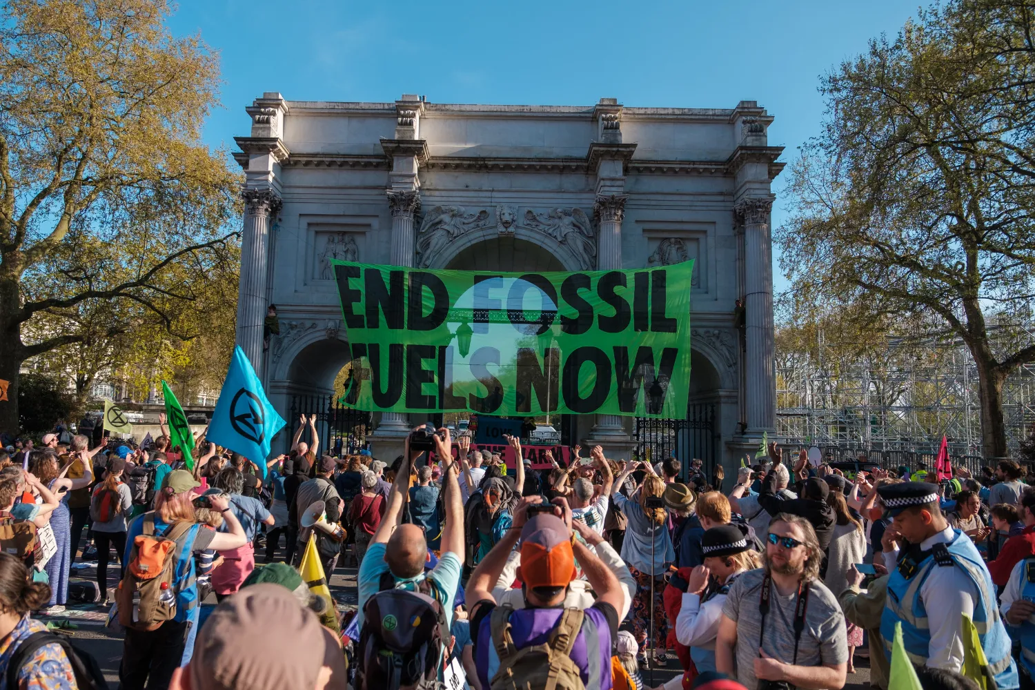 Manifestation climatique devant un arc monumental, avec une grande banderole verte portant le message 'End Fossil Fuels Now'