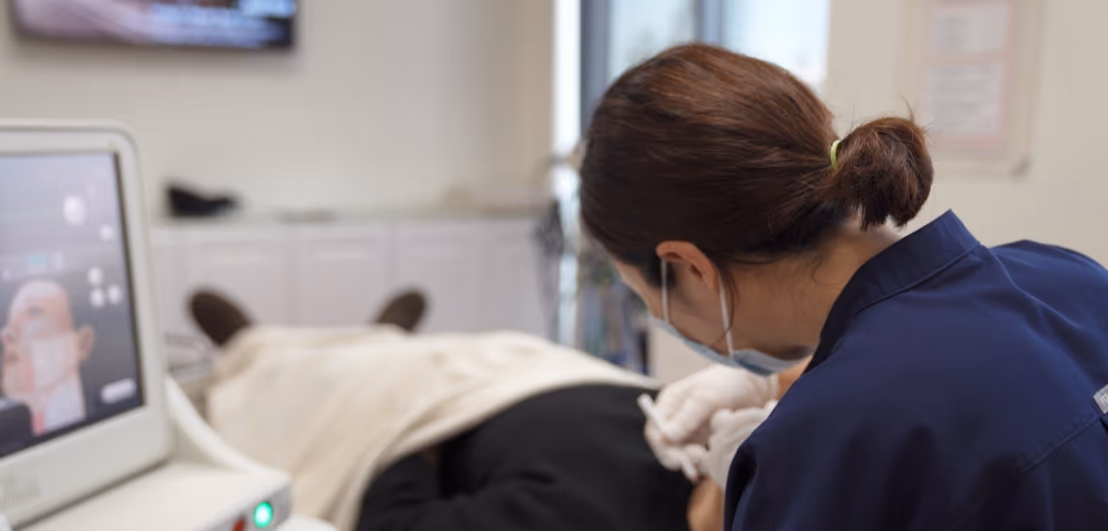 Dr Kim's assistant medical professional wearing gloves and a mask administering an injection to a patient lying down covered with a blanket.