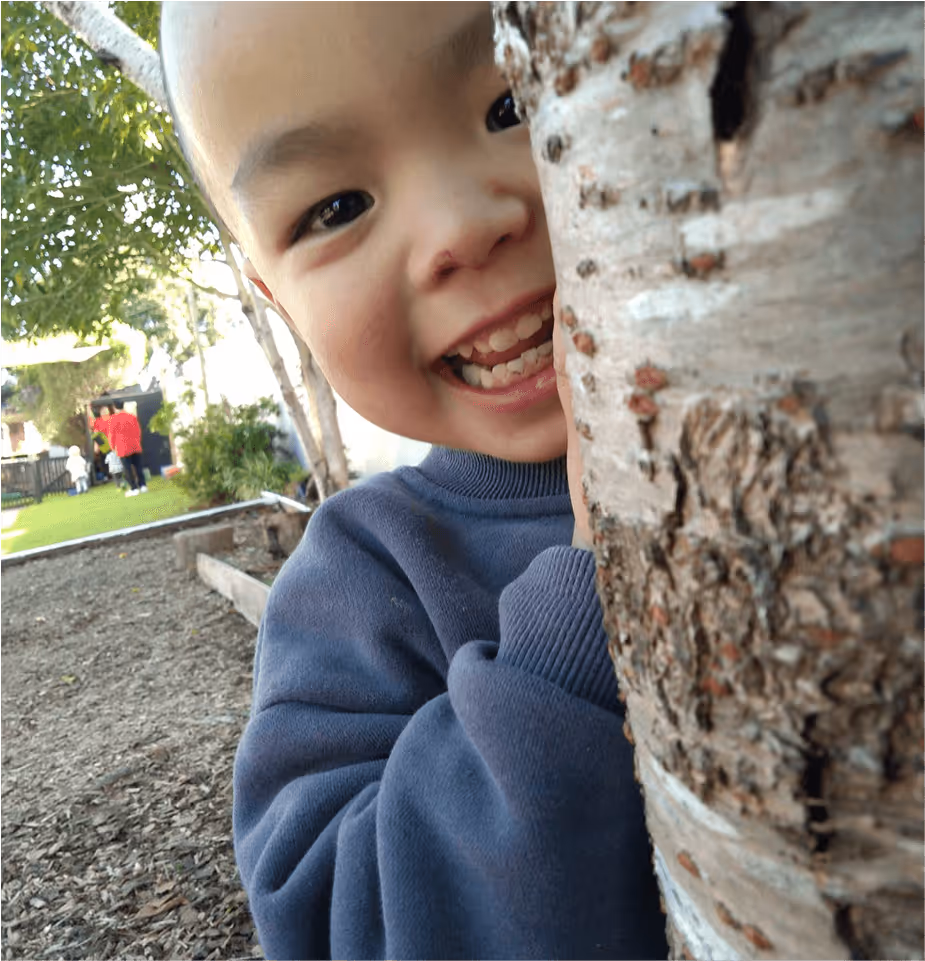 A young child wearing a blue sweater peeks playfully from behind a tree trunk, smiling brightly in an outdoor setting.