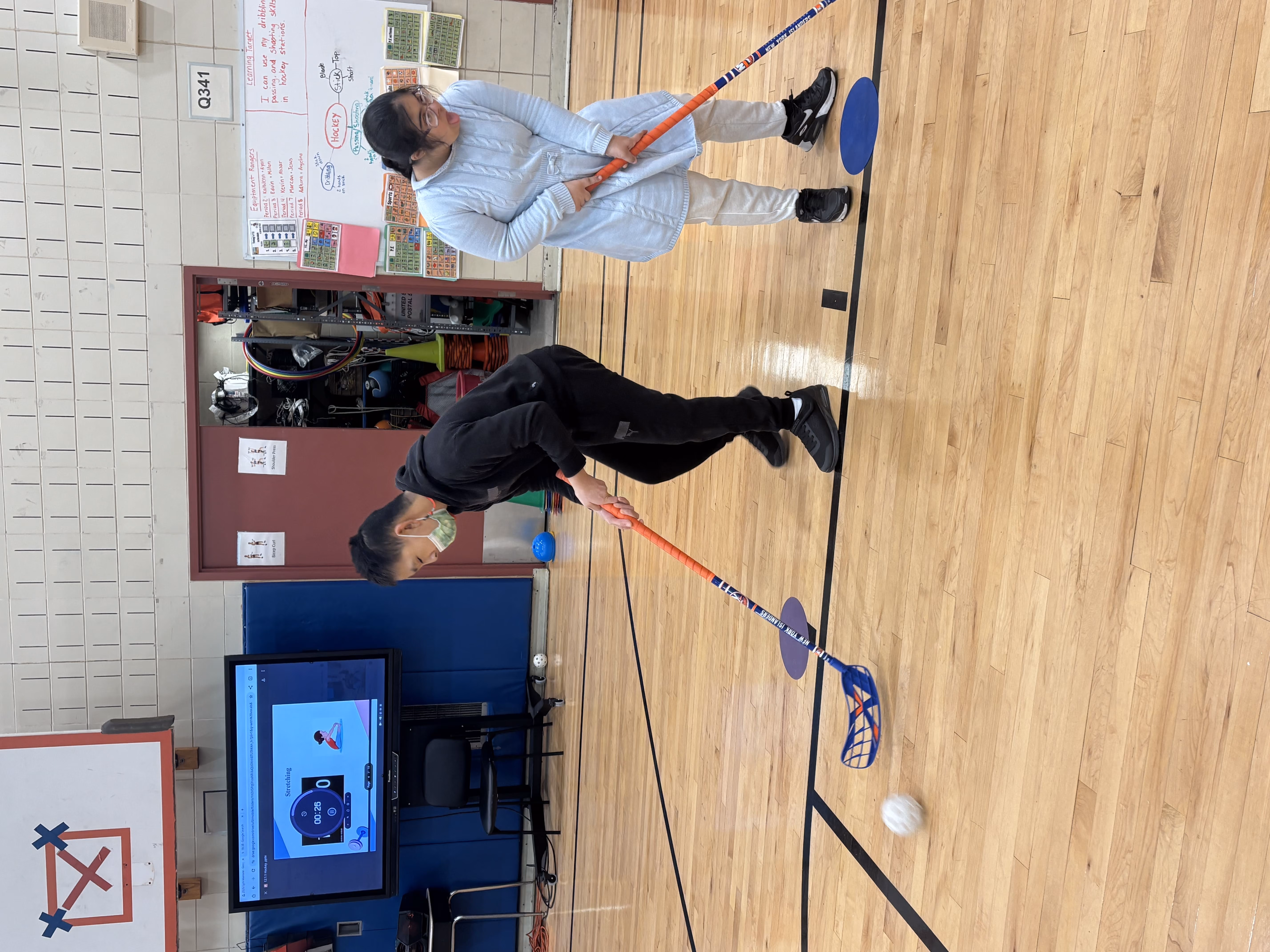 Two students playing floor hockey with sticks in school gymnasium