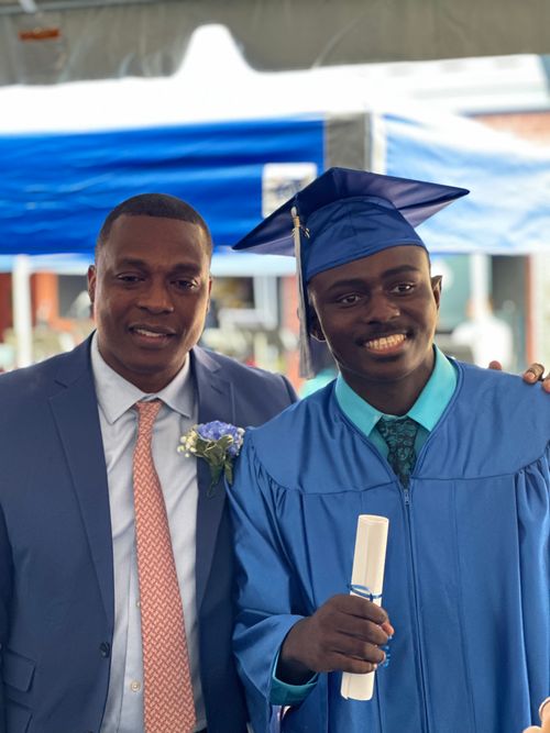 Graduate in blue cap and gown holding diploma poses with adult