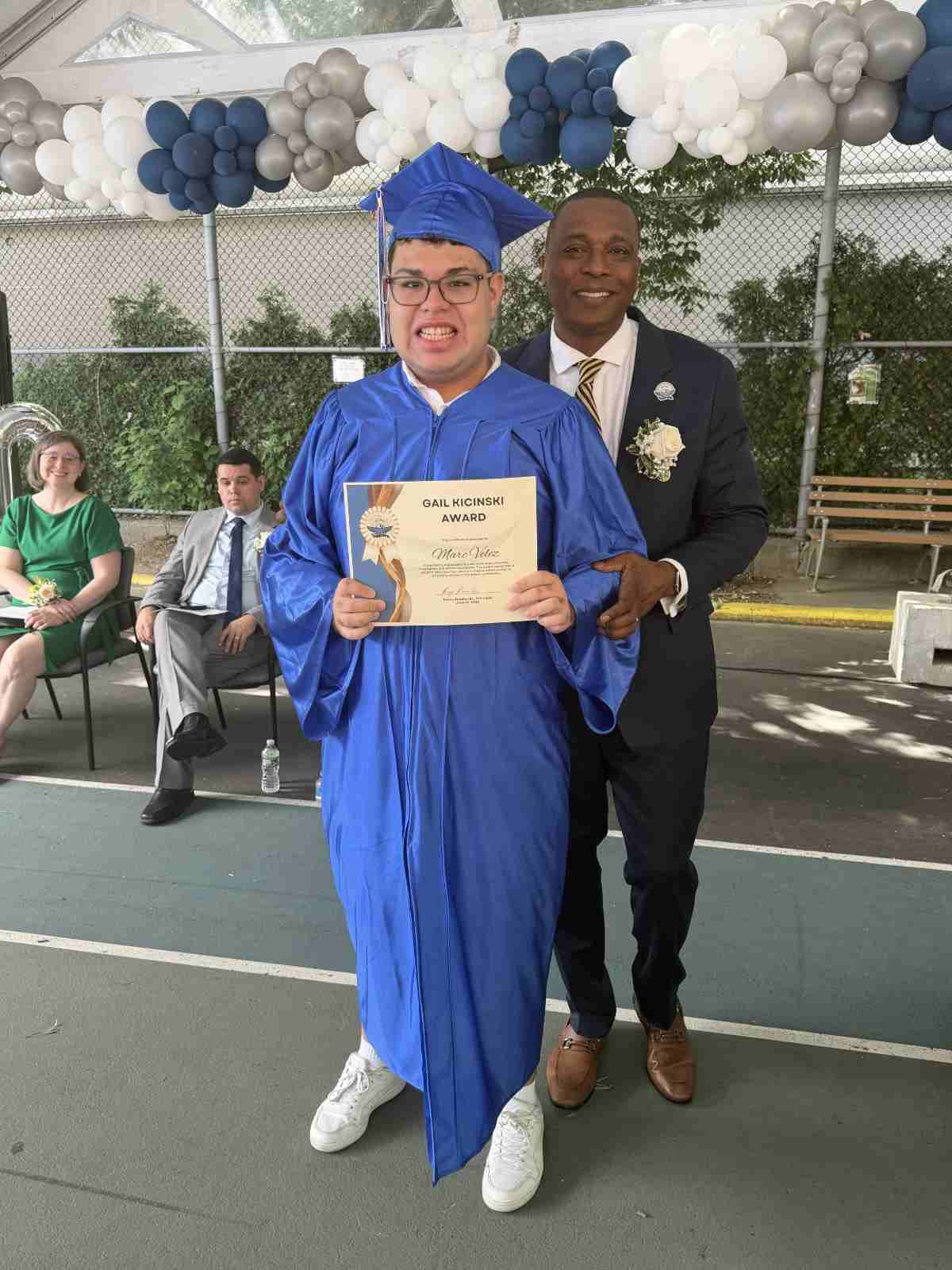 Graduate in blue cap and gown poses with three adults outdoors