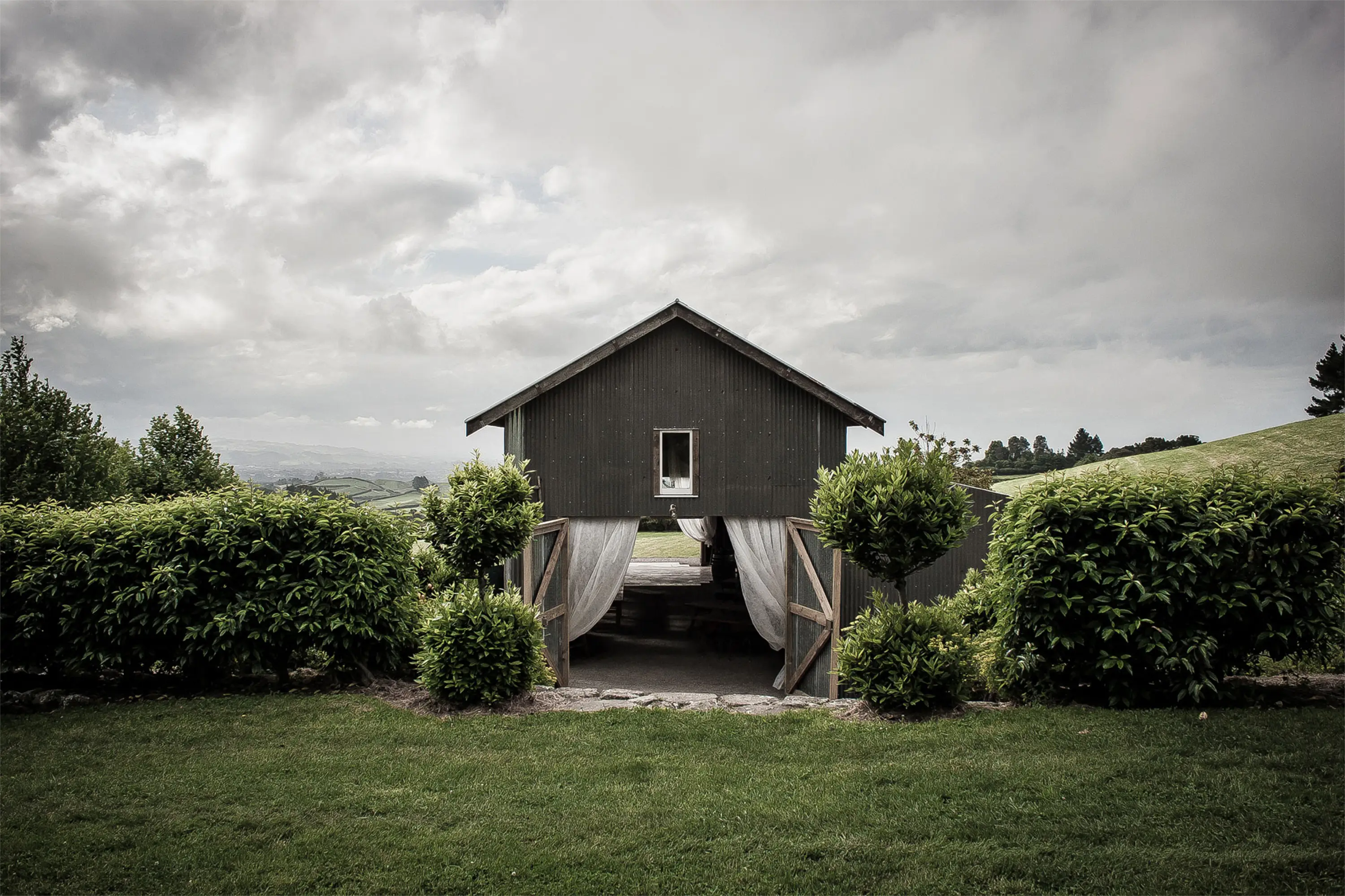 A small black wooden barn with open doors sits between neatly trimmed green bushes, surrounded by grass and rolling hills under a cloudy sky.