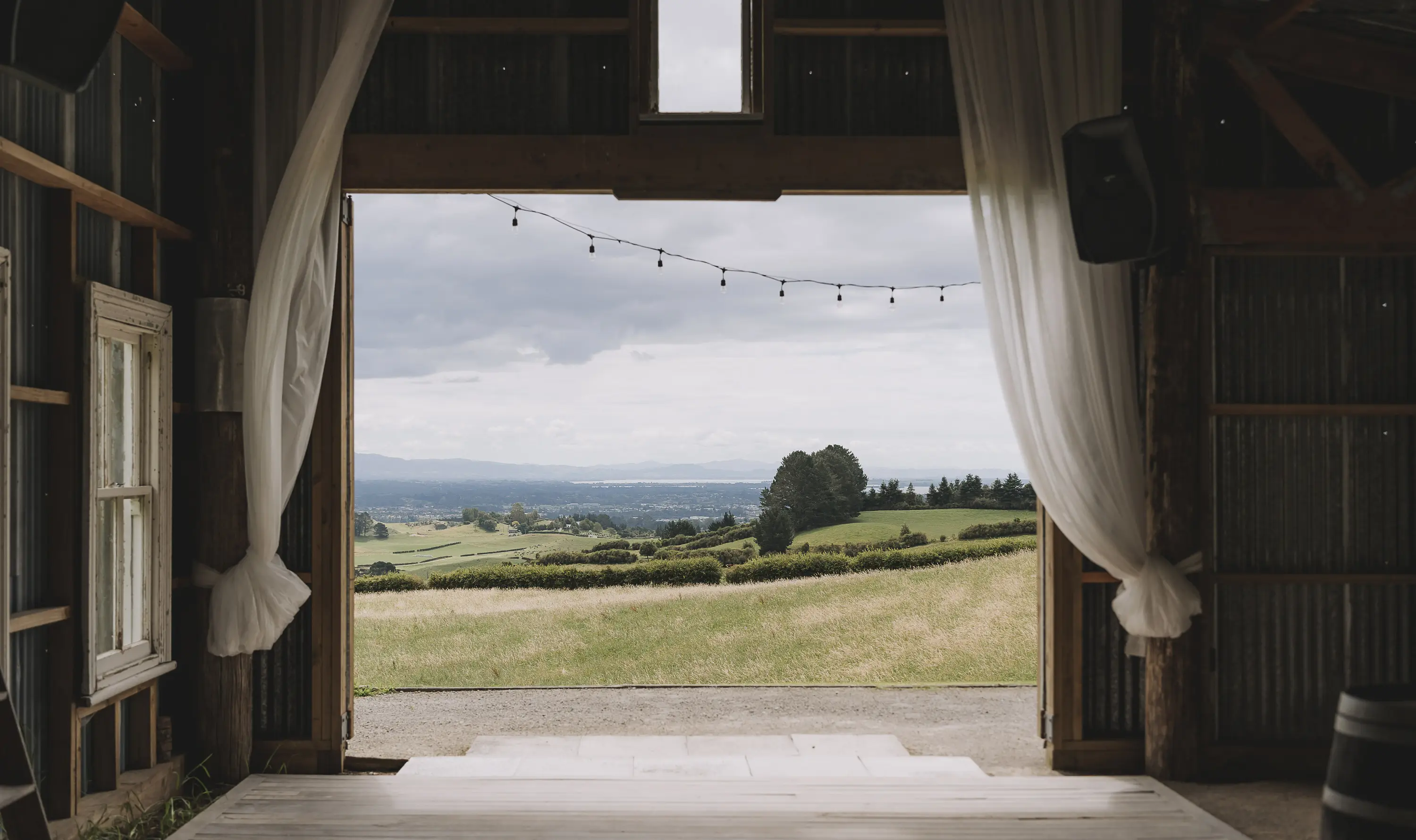 View of rolling green hills and distant mountains seen through the large open doorway of a rustic barn with white curtains and string lights, under a cloudy sky.