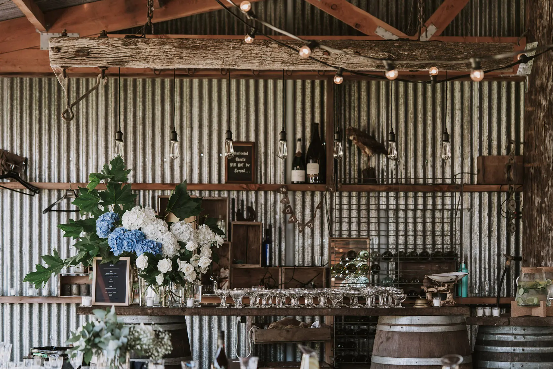 A rustic bar with a corrugated metal wall, shelves holding bottles and glassware, hanging string lights, flower arrangements with white and blue blooms, and wooden barrels used as decor.
