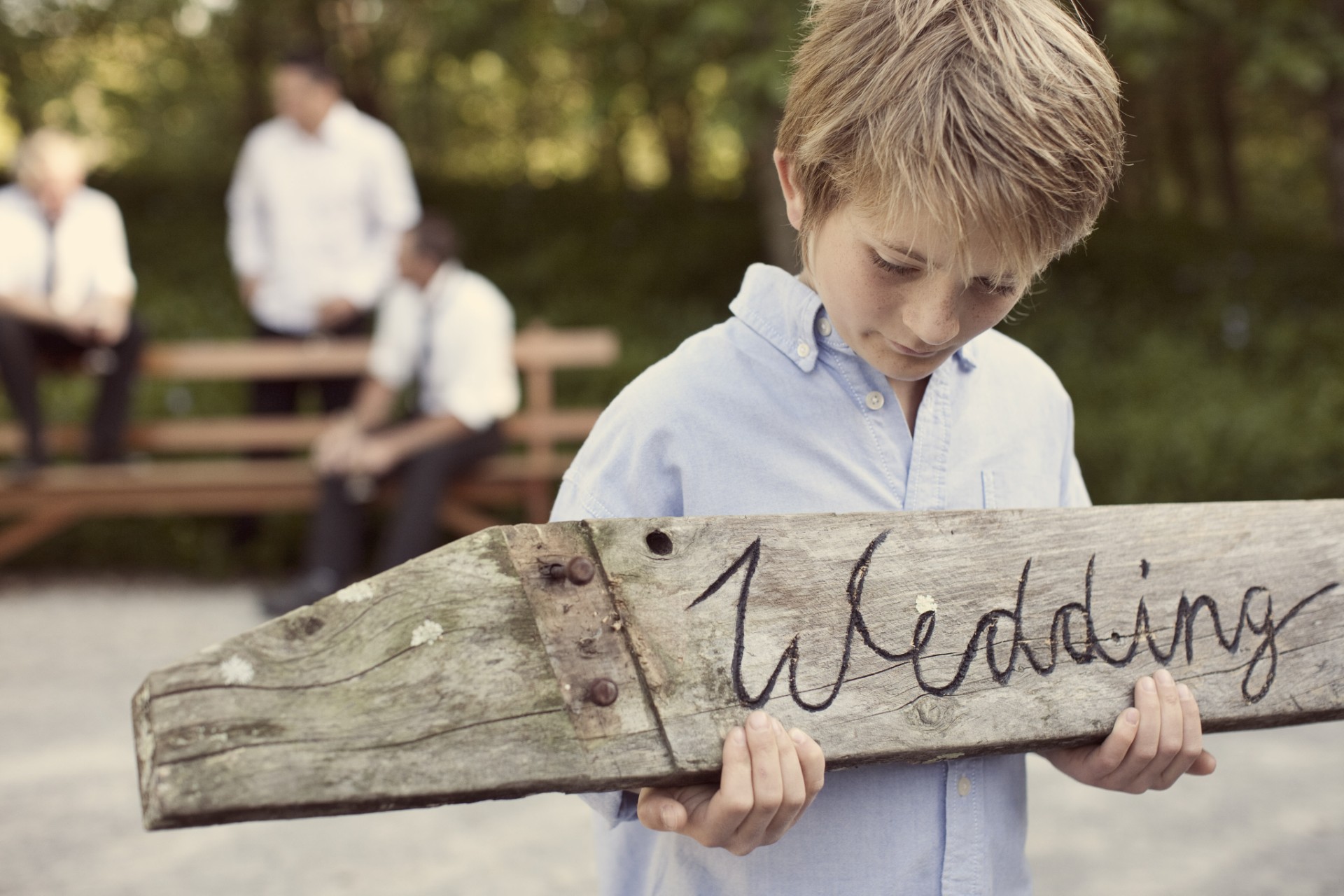 A young boy in a light blue shirt holds a rustic wooden sign that says "Wedding." In the blurred background, three people in white shirts sit on a bench outdoors.