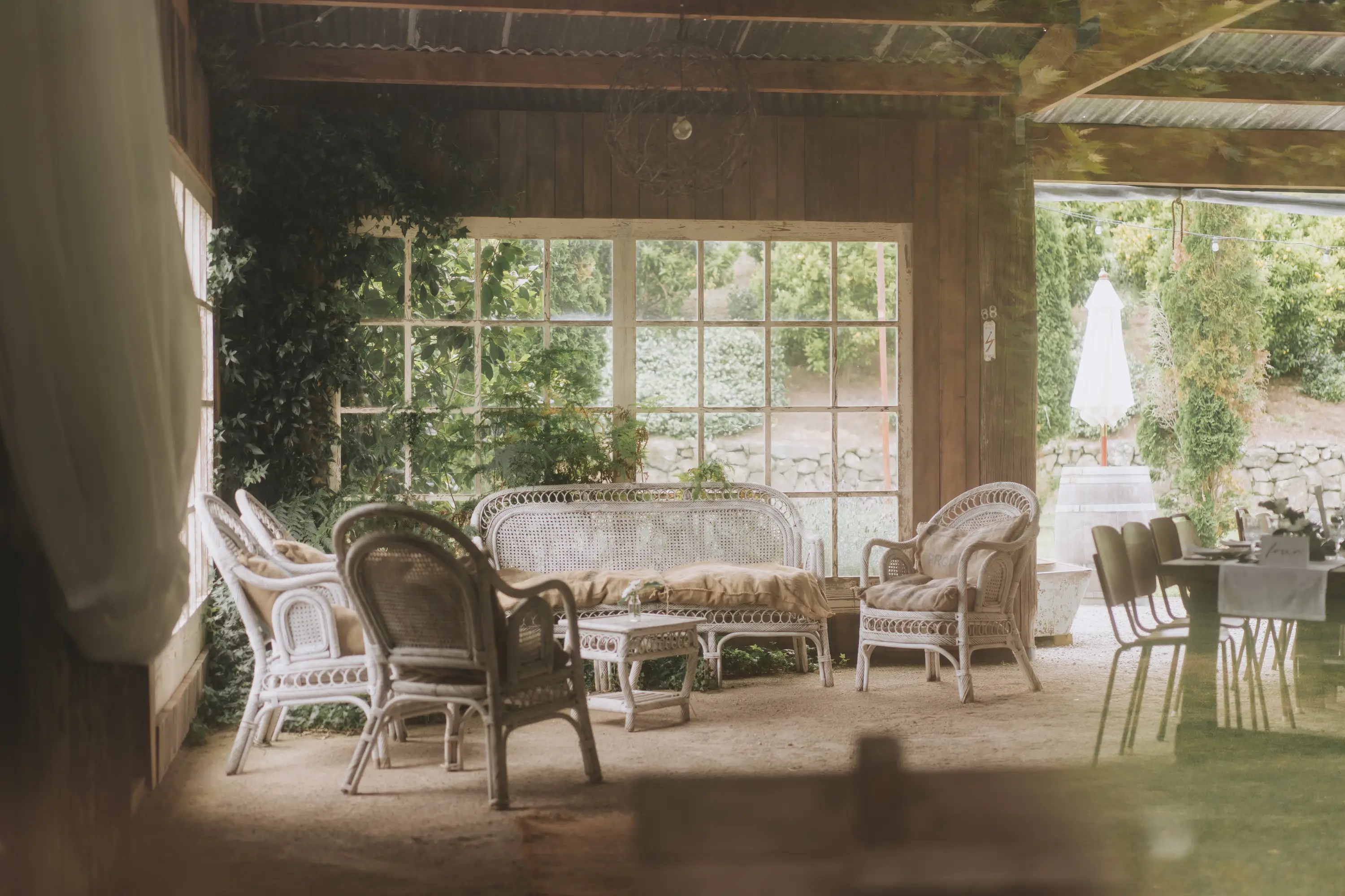 A cozy indoor seating area with wicker chairs and a sofa, surrounded by large windows that reveal greenery outside. Sunlight filters in, creating a warm and inviting atmosphere.