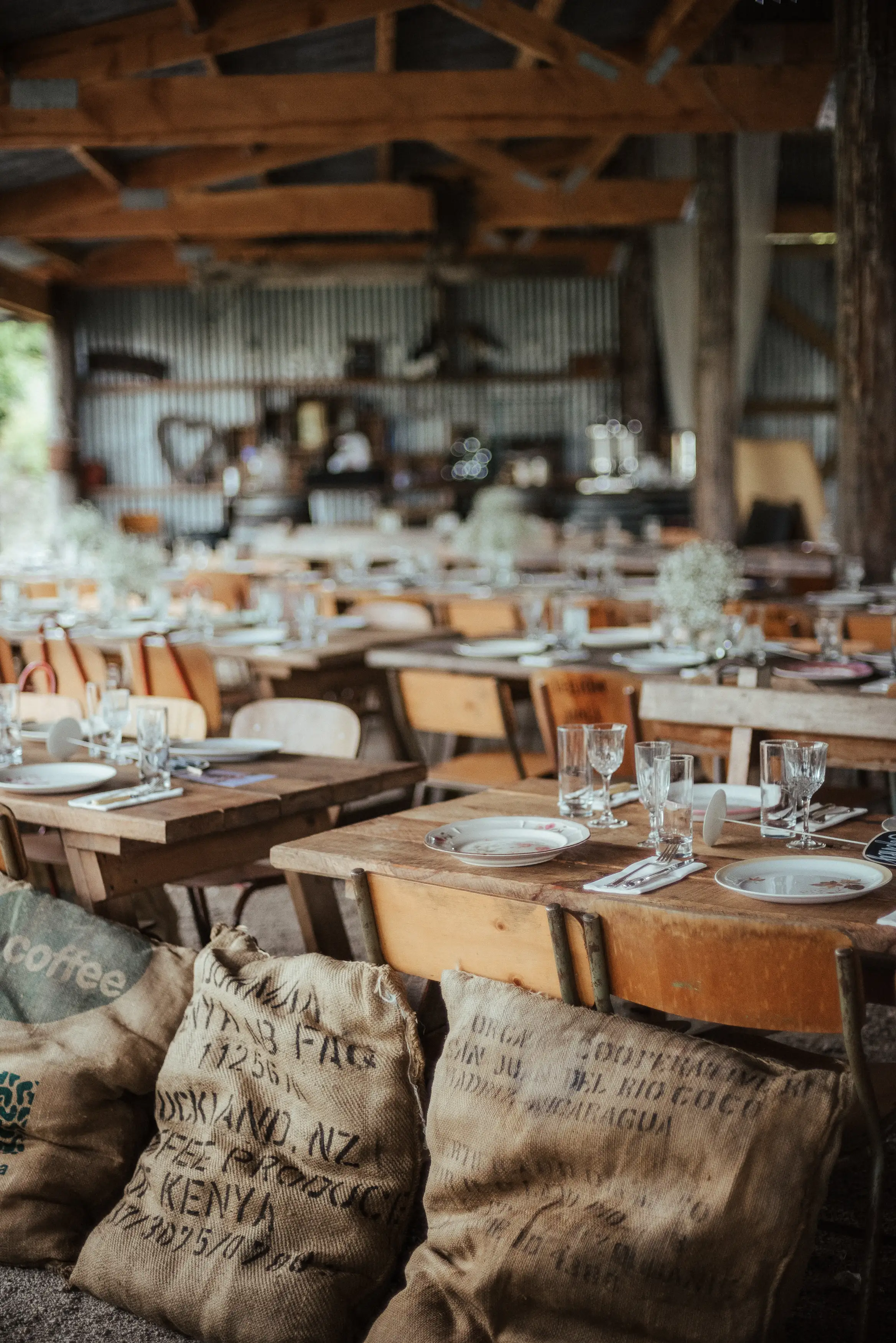 Rustic event setup with wooden tables and chairs arranged in rows, set with plates, glasses, and cutlery. Burlap coffee sacks are stacked in the foreground, and the venue features exposed beams and corrugated metal walls.