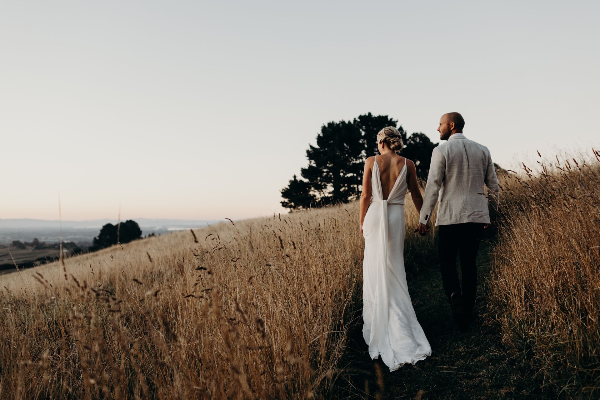 A bride and groom walk hand in hand through a grassy field at sunset, with tall golden grass and trees in the background. The bride wears a white dress and the groom wears a light jacket and dark pants.