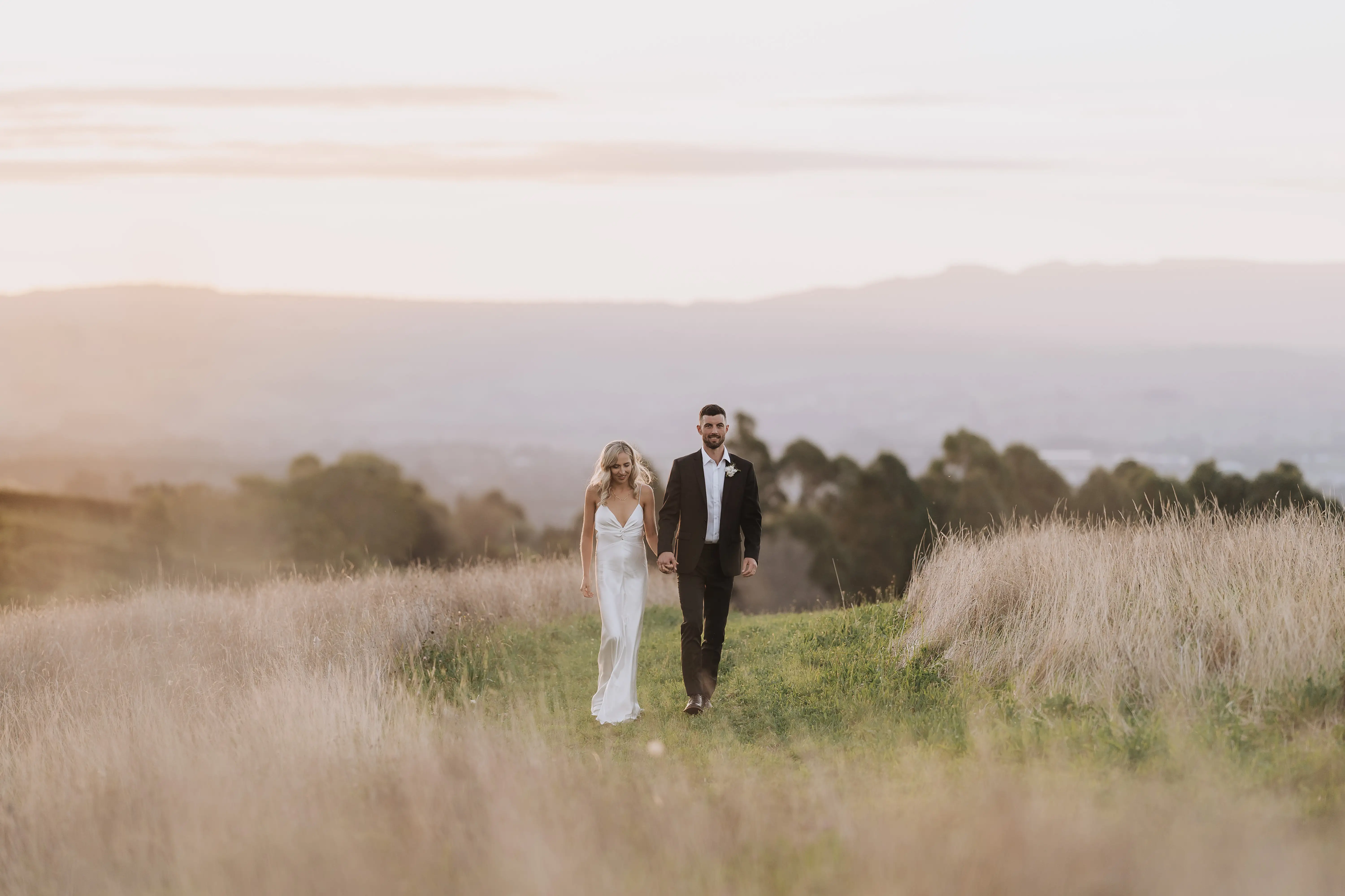 A bride in a white dress and a groom in a dark suit walk hand in hand along a grassy path surrounded by golden fields at sunset, with soft hills visible in the distance.