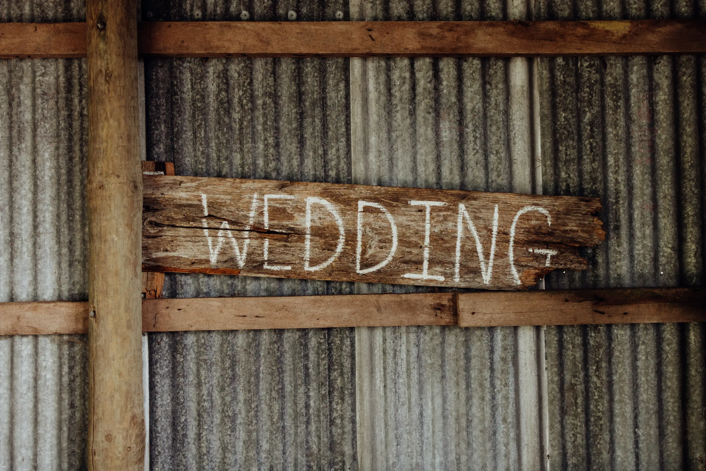 A rustic wooden sign with the word "WEDDING" painted in white, hanging on a corrugated metal wall with exposed wooden beams.