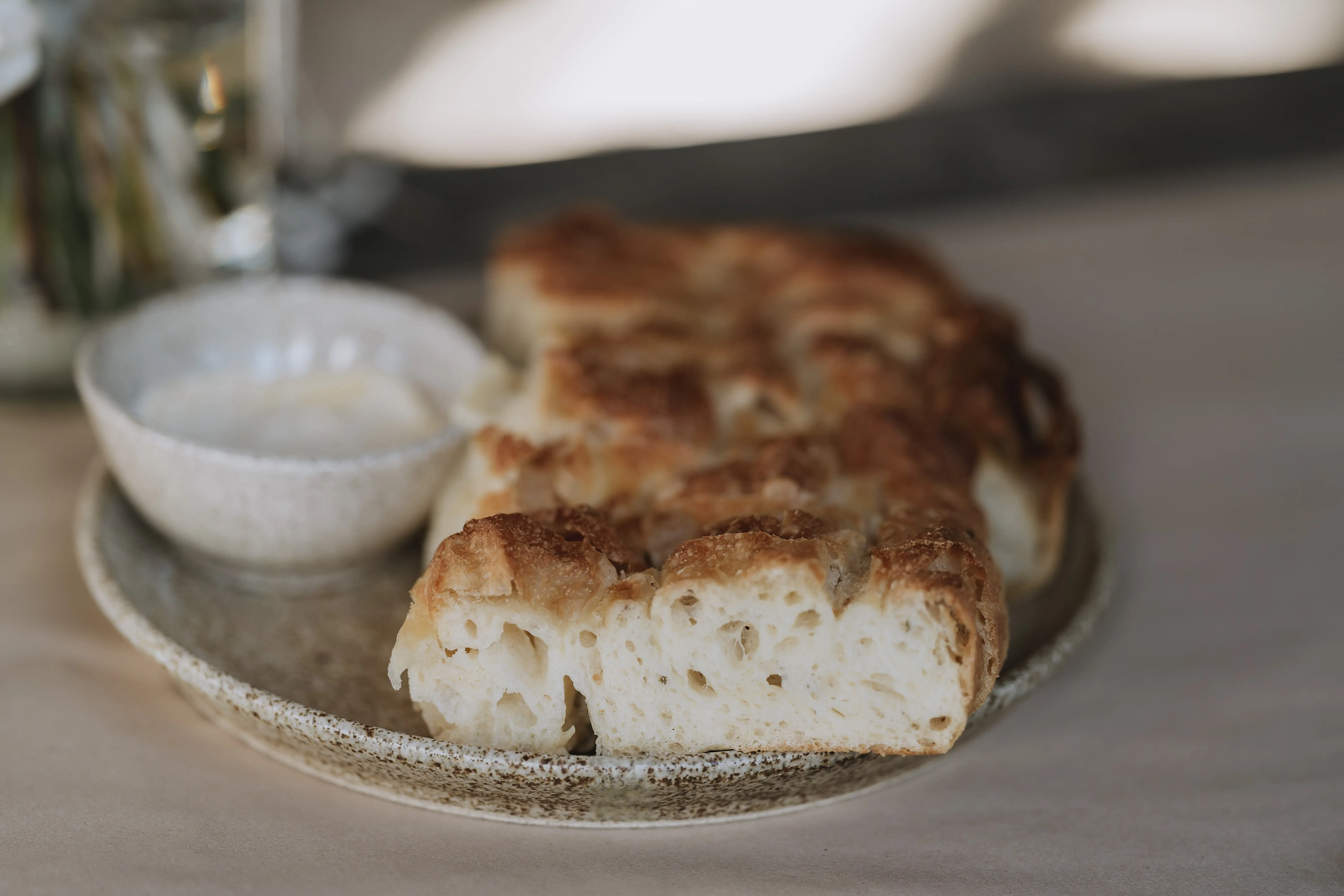 A plate with sliced pieces of focaccia bread next to a small bowl of creamy dipping sauce on a light-colored table.