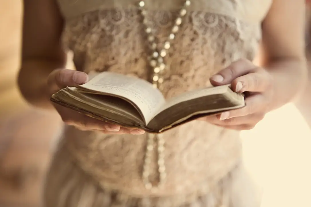 A person wearing a lace dress and a long beaded necklace holds an open book with both hands, reading. The image is softly focused, creating a warm, vintage atmosphere.