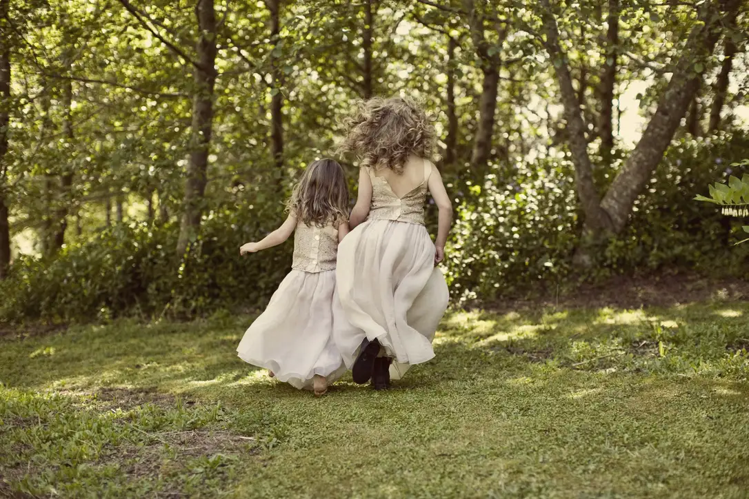 Two young girls wearing light-colored dresses run barefoot through a grassy area surrounded by trees, with sunlight streaming through the leaves, creating a serene and playful outdoor scene.